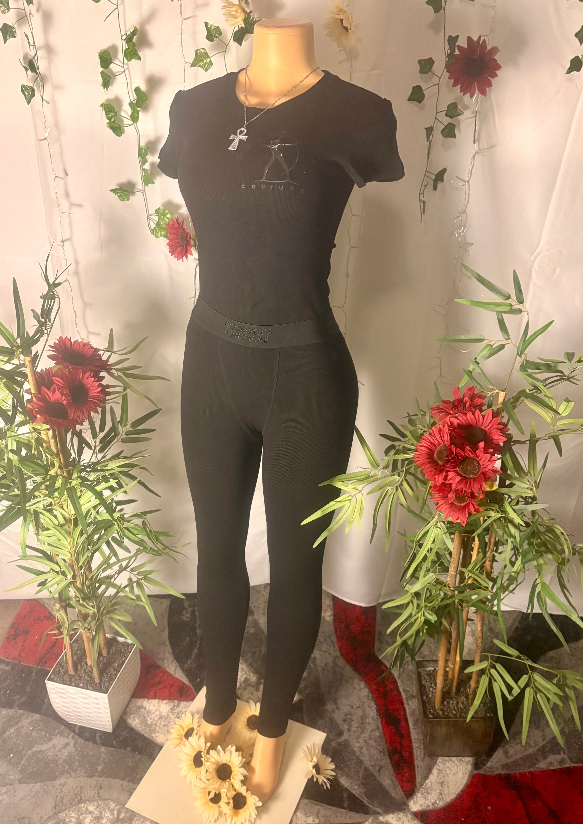 Mannequin wearing Delisha's Couture branded with logo midnight black colored ribbed textured two-piece pants outfit with a decorative background of plants and flowers.