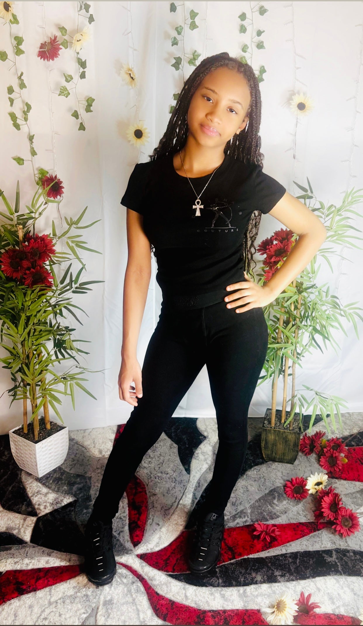 Little diva modeling Delisha's Couture branded midnight black ribbed textured two-piece pants outfit with a decorative background featuring plants and flowers.