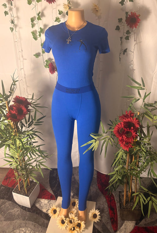 Mannequin wearing Delisha's Couture branded with logo, ribbed textured two-piece magnetic blue outfit with decorative plants and flowers in the background
