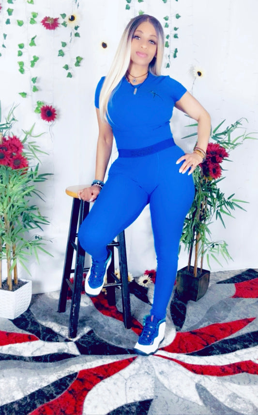 Model wearing Delisha's Couture branded with logo magnetic blue ribbed two-piece outfit propped up on a stool with floral and decorative elements in the background