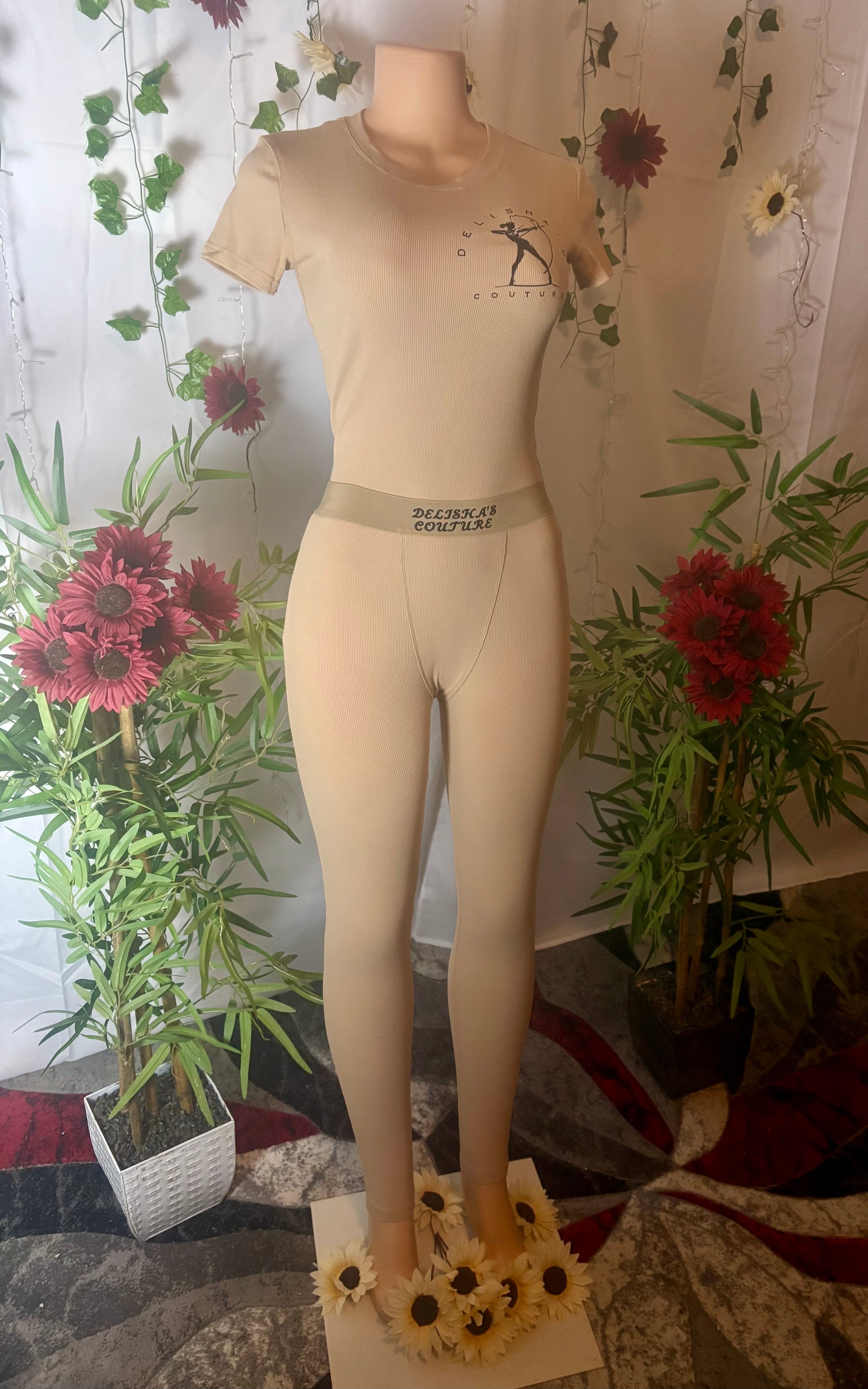Mannequin wearing Delisha's Couture branded with logo ribbed textured lucky beige two-piece pants outfit with floral decorations and plants in the background